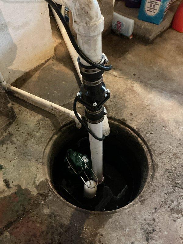 Responded to flooded basement in 1950s-era home with suspected sump pump failure. Upon inspection, found existing green submersible pump in concrete sump pit with standing water and aging discharge system. Assessed pump functionality, verified electrical connections, and inspected white PVC discharge piping secured with metal hose clamps. Addressed water accumulation issue, tested pump operation, and confirmed proper drainage. System restored to working order with pump successfully removing standing water from basin.