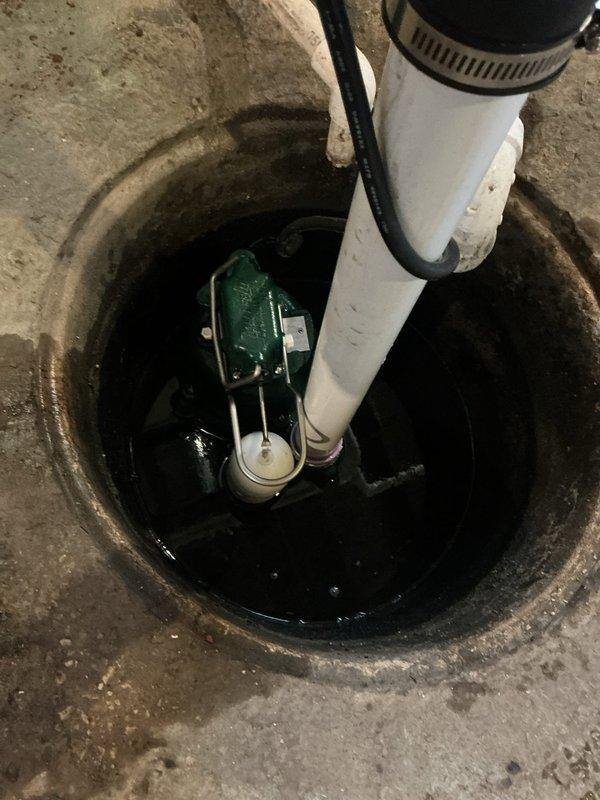 Responded to flooded basement in 1950s-era home with suspected sump pump failure. Upon inspection, found existing green submersible pump in concrete sump pit with standing water and aging discharge system. Assessed pump functionality, verified electrical connections, and inspected white PVC discharge piping secured with metal hose clamps. Addressed water accumulation issue, tested pump operation, and confirmed proper drainage. System restored to working order with pump successfully removing standing water from basin.