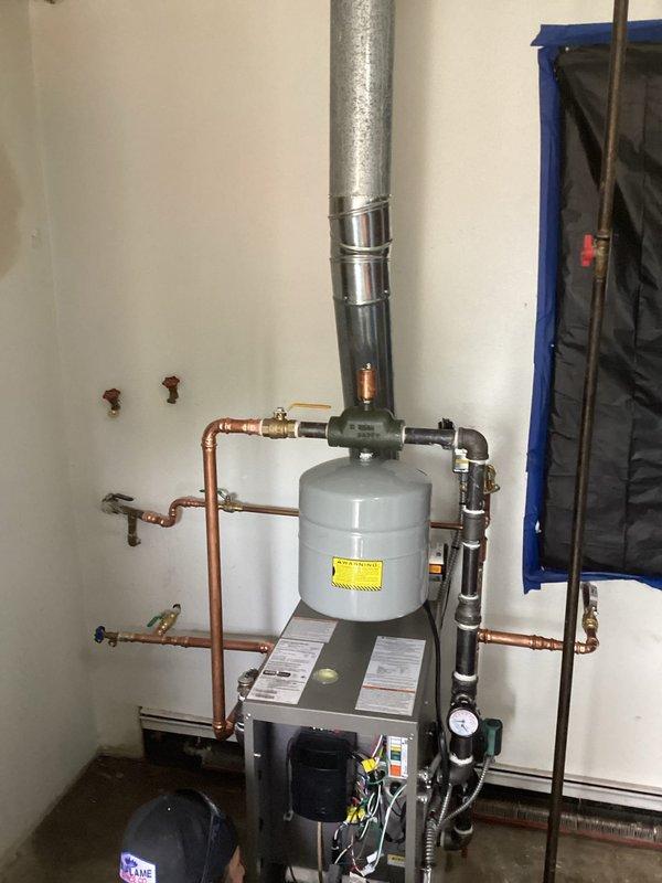Completed installation of floor-mount gas-fired boiler system in basement utility area, accessing through rear entrance as specified. System includes new boiler unit with integrated expansion tank, copper supply and return piping with brass fittings and isolation valves, black PVC venting with pressure gauge, and proper connections to existing hydronic heating distribution system. All components installed per manufacturer specifications with appropriate clearances, system tested and operational.