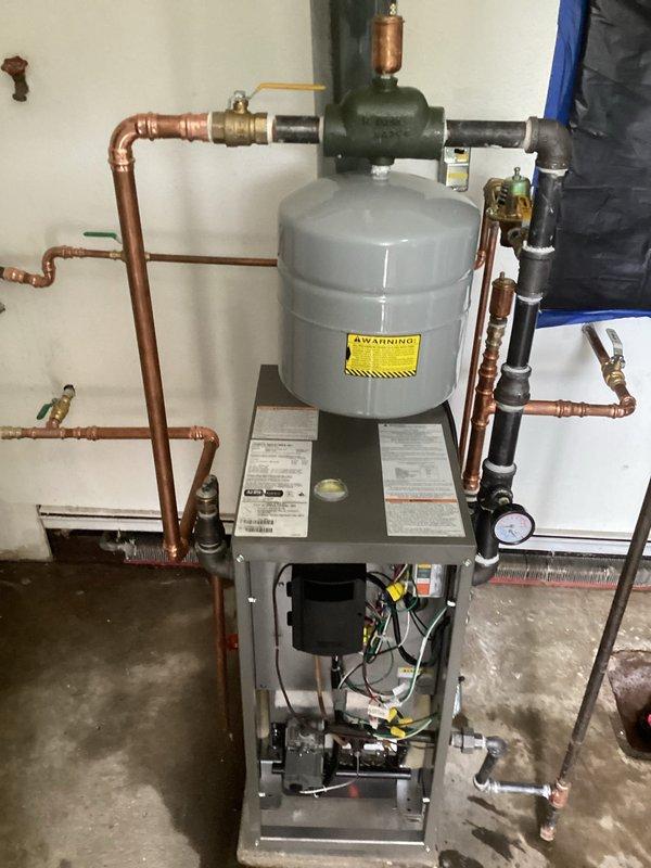 Completed installation of floor-mount gas-fired boiler system in basement utility area, accessing through rear entrance as specified. System includes new boiler unit with integrated expansion tank, copper supply and return piping with brass fittings and isolation valves, black PVC venting with pressure gauge, and proper connections to existing hydronic heating distribution system. All components installed per manufacturer specifications with appropriate clearances, system tested and operational.