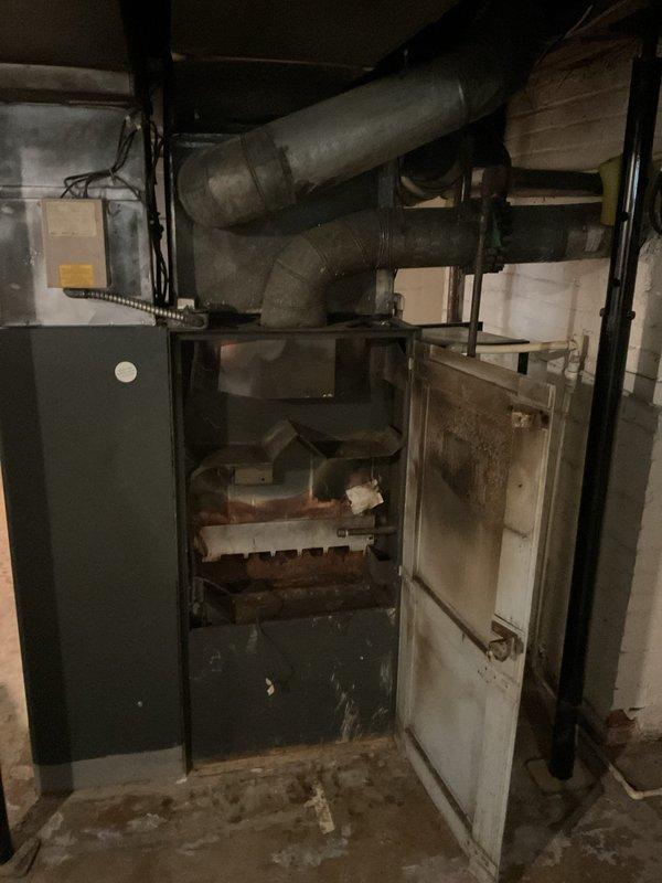 Completed furnace replacement estimate for aged HVAC system in basement utility area. Assessed existing dark-colored heating unit with deteriorated ductwork showing significant dust accumulation and wear. Evaluated green metal cabinet enclosure exhibiting rust damage and moisture exposure on door panels. Photographed current equipment configuration including flexible metallic conduit connections, horizontal metal ductwork, and mechanical components within open compartment showing aged metal surfaces. Documented basement conditions including concrete floor with moisture staining and overall equipment deterioration indicating need for system replacement.