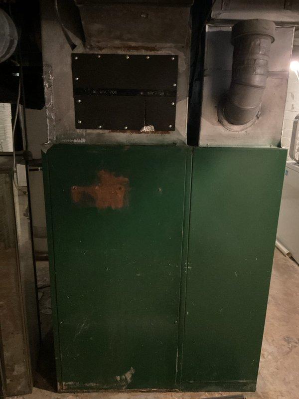 Completed furnace replacement estimate for aged HVAC system in basement utility area. Assessed existing dark-colored heating unit with deteriorated ductwork showing significant dust accumulation and wear. Evaluated green metal cabinet enclosure exhibiting rust damage and moisture exposure on door panels. Photographed current equipment configuration including flexible metallic conduit connections, horizontal metal ductwork, and mechanical components within open compartment showing aged metal surfaces. Documented basement conditions including concrete floor with moisture staining and overall equipment deterioration indicating need for system replacement.