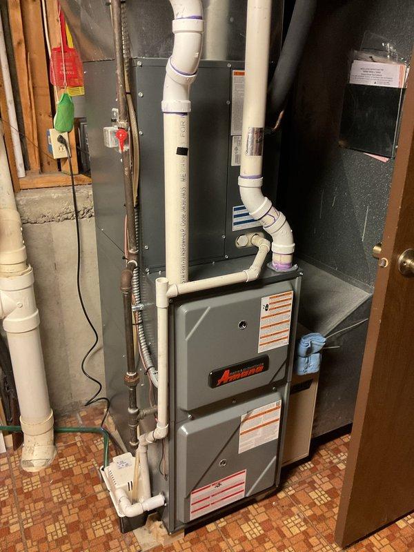 Completed equipment estimate for HVAC system replacement including indoor Amana furnace unit installed in basement utility area and outdoor air conditioning condenser unit positioned on concrete pad against exterior wall. Installation includes proper PVC drainage and ventilation piping, insulated refrigerant lines, metal ductwork integration, electrical disconnect box, and all necessary connections for complete heating and cooling system operation.