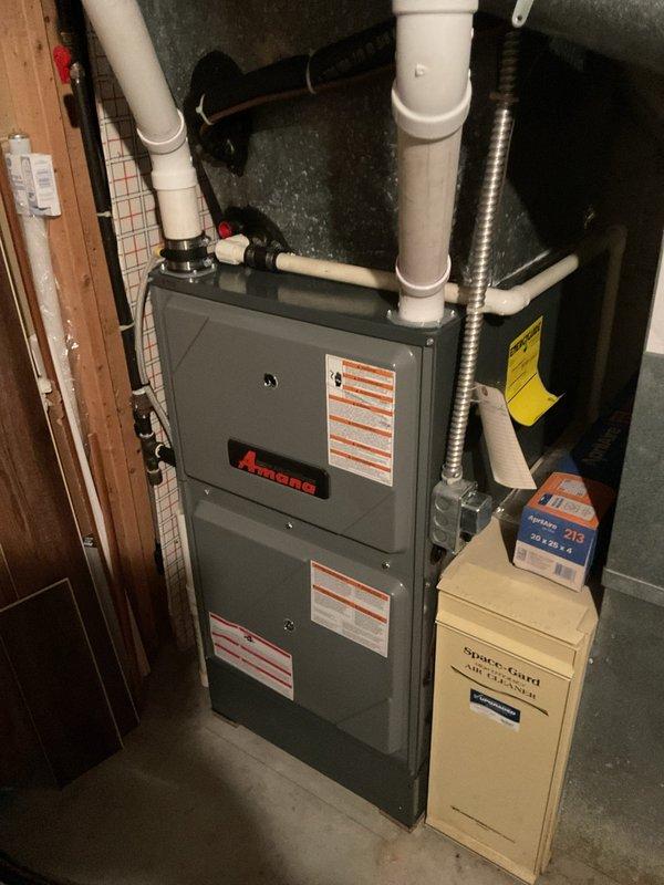 Conducted comprehensive site evaluation of existing HVAC system for complete replacement estimate. Assessed outdoor Amana condenser unit positioned on concrete pad in residential backyard, noting equipment age, condition, and accessibility for replacement. Evaluated indoor Amana furnace and air handler configuration in basement utility area, documenting current system specifications, venting arrangement with PVC exhaust pipes, and ductwork connections. Photographed both outdoor and indoor equipment to document existing installation parameters, clearances, and spatial considerations necessary for accurate replacement system sizing and installation planning.