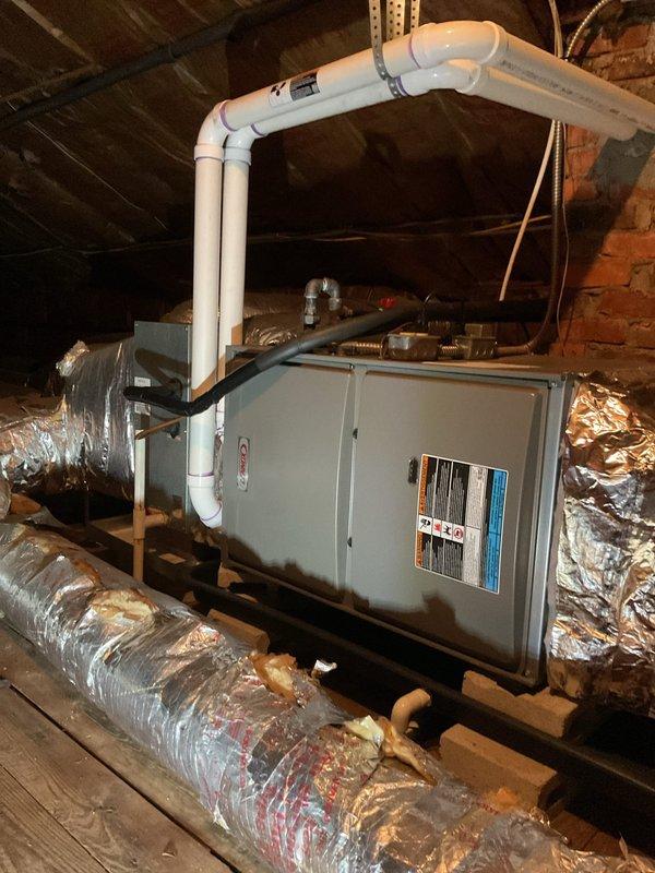 Conducted comprehensive HVAC system evaluation in attic space for equipment replacement estimate. Assessed existing gray metal air handler unit mounted on wooden support beams, including inspection of white PVC condensate drain line configuration with multiple elbows running along ceiling joists. Evaluated flexible metallic ductwork on both sides of unit and photographed manufacturer specification labels for equipment sizing and compatibility verification. Documented attic access conditions, storage items, and workspace constraints for replacement planning purposes.