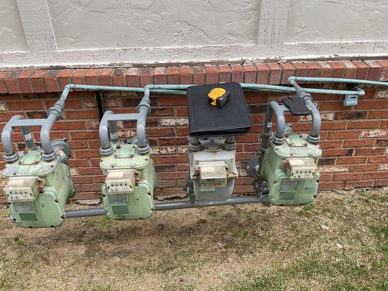 Completed installation site visit and documentation for multi-unit residential HVAC system. Installed interior wall-mounted access panel with integrated ventilation grille for system access and air circulation. Panel features secure mounting with appropriate fasteners and louvers for proper airflow management. Documented existing natural gas meter bank serving multiple units, including four analog meters with associated PVC piping network, central junction box, and safety shutoff valve. All installations photographed for service records and quality assurance purposes.