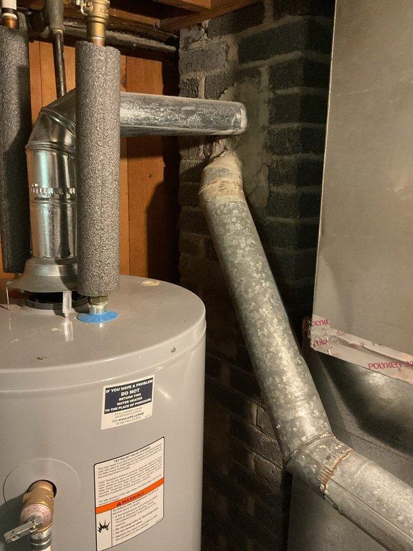 Performed comprehensive assessment of existing HVAC system for complete air conditioning replacement. Evaluated outdoor condenser unit with cylindrical design and black horizontal grille fins currently positioned on grass adjacent to building exterior. Documented existing electrical disconnect box and utility connections, including conduit routing from building-mounted components to ground-level equipment. Assessed indoor components including water heater and associated plumbing infrastructure in basement utility area to ensure compatibility with proposed AC system replacement and verify adequate clearances for new equipment installation.