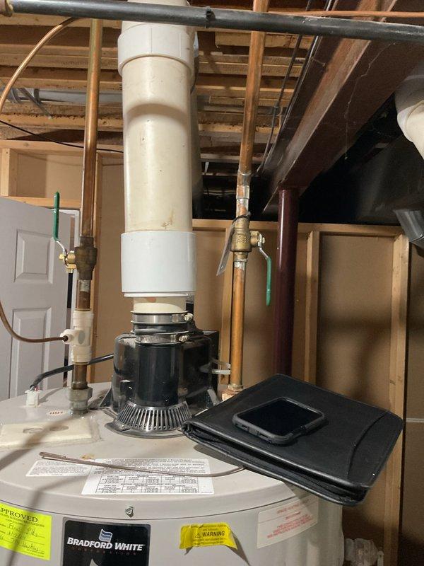 Completed comprehensive assessment of existing HVAC system and water heating equipment for replacement estimate. Evaluated current Bradford White water heater installation located in unfinished basement utility area, documenting existing configuration including power-vented exhaust system with black cylindrical draft inducer assembly mounted on top of unit. Inspected all plumbing connections including copper supply lines, brass shut-off valves, green-handled isolation valve, and flexible tubing connections. Assessed current PVC venting configuration running vertically through ceiling joists. Photographed existing installation showing water heater positioning beneath exposed floor framing, connection points, safety labels, and surrounding utility space constraints. Examined furnace system requiring replacement and documented spatial requirements for new equipment installation. Provided detailed replacement recommendations for both heating system and water heater upgrade, including equip