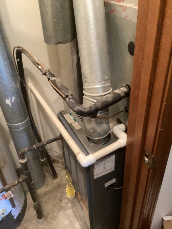Completed comprehensive system evaluation for full HVAC replacement at apartment complex mechanical room. Documented existing residential heating and cooling equipment including dark-cased furnace unit with white PVC exhaust venting, extensive galvanized metal ductwork distribution system with vertical and horizontal runs, and adjacent water heater with copper and flexible supply lines. Assessed vertical aluminum duct installation secured with metal clamps running through confined utility space with exposed insulation and framing. Photographed all major components including furnace specifications plate, duct connections with metal collars and clamps, PVC condensate and venting systems, and mechanical room layout to provide accurate replacement estimate for aging equipment in basement utility area.