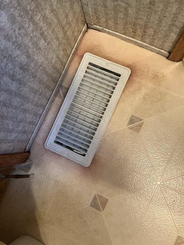 Responded to customer complaint of scorching smell from furnace register. Upon inspection, discovered significant heat damage and discoloration around floor register and ductwork, with visible staining and debris accumulation in the vent opening. Documented conditions with photographs showing brown discoloration pattern around metallic duct opening and compromised floor register, indicating potential airflow restriction or overheating issue requiring further system evaluation.