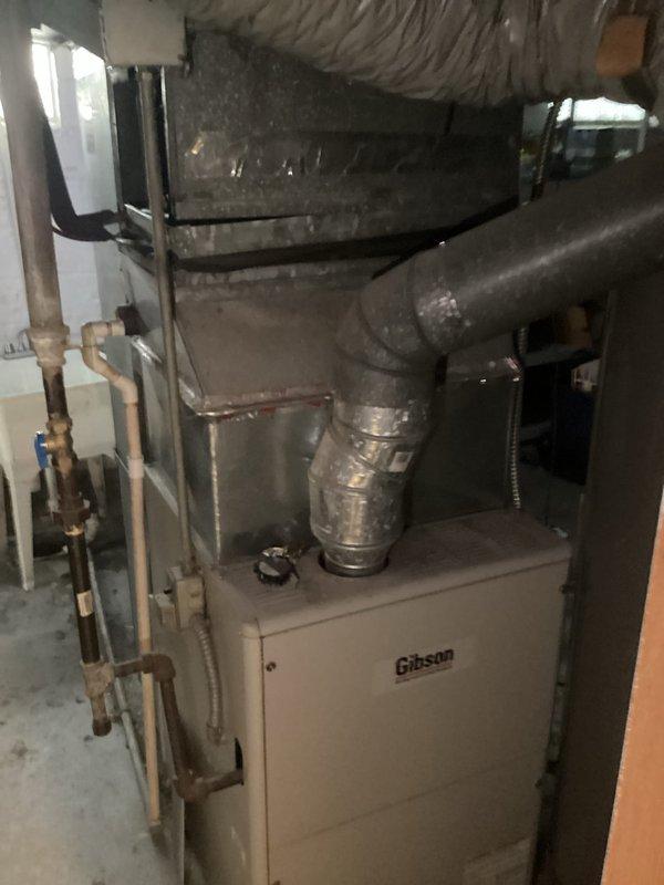 Conducted comprehensive site assessment for complete HVAC system replacement at residential rental property. Evaluated existing outdoor condensing unit with protective cage enclosure positioned on concrete pad adjacent to building foundation, noting electrical service connections and meter configuration. Inspected basement mechanical room containing Gibson furnace with associated ductwork, including flexible insulated ducting and rigid metal supply/return piping. Documented current system layout, electrical infrastructure, refrigerant line routing, and existing copper plumbing connections to provide accurate replacement specifications and equipment sizing recommendations for property management consideration.