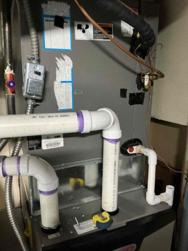 Documented existing HVAC system installation for replacement estimate purposes. The current setup includes a gray metal furnace unit with associated condensate drainage system, electrical connections, and refrigerant lines. Photographed all components including PVC drain piping with proper trap configuration, flexible electrical conduit, and existing cabinet assembly to provide comprehensive documentation for preparing replacement equipment quote and identifying any modifications needed for new system installation.