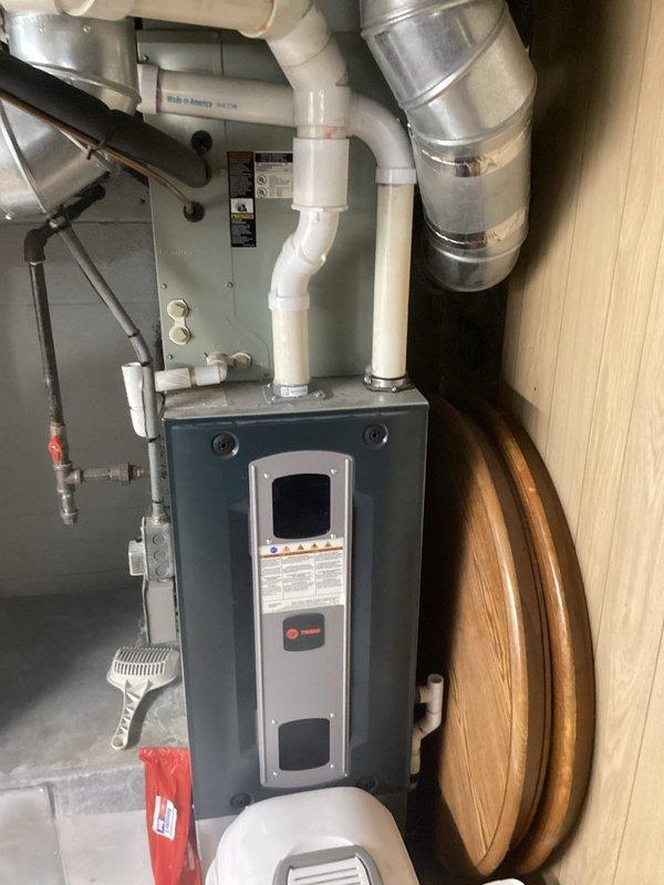 Completed full HVAC system replacement including outdoor Trane condenser unit and indoor furnace with air handler. The new condenser was installed on existing concrete pad against exterior wall with proper clearances maintained, refrigerant lines were run and insulated, and all electrical connections completed. Indoor installation included new furnace with viewing window, wall-mounted air handler, PVC condensate drain lines, humidifier unit, and metallic supply ductwork with all components properly integrated and tested for operation.