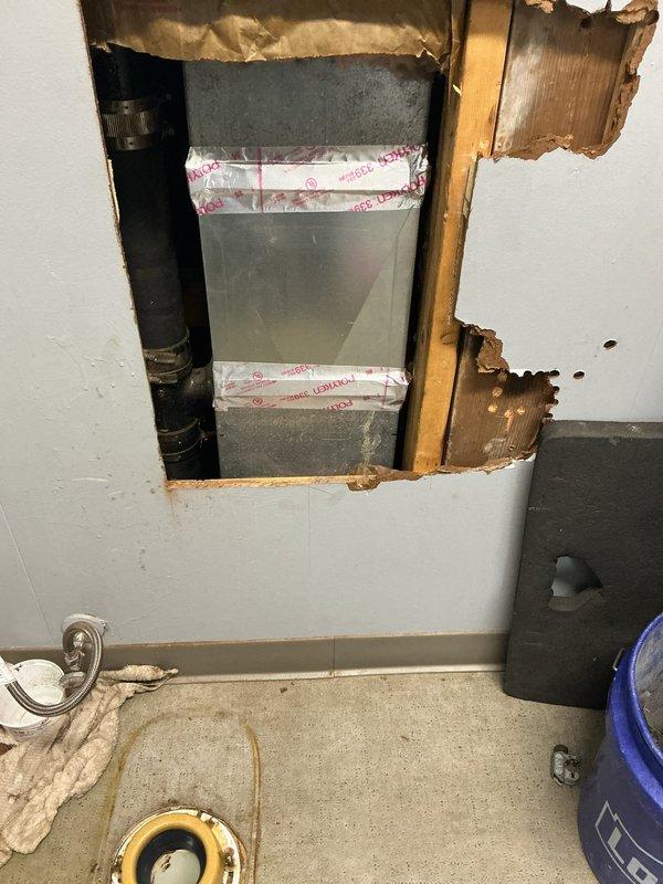 Accessed plumbing system through wall cavity between toilet and pedestal sink to address water damage and deterioration. Removed section of drywall to expose wooden framing studs and assess extent of damage to interior wall materials. Located and inspected black vertical drain pipe and metal ductwork within wall cavity. Removed and reinstalled affected plumbing components to resolve issue. Documented existing water staining and structural deterioration for repair assessment.