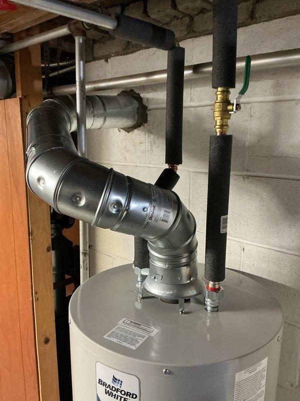 Conducted on-site assessment of Bradford White tanked water heater installation in basement utility area. Evaluated existing water heater system including venting configuration, piping connections, and overall installation quality. Verified proper placement and leveling of unit against concrete block foundation wall. Inspected metallic vent pipe system with multiple elbows and joints for proper exhaust routing. Examined plumbing connections including foam-insulated supply lines, brass valving with control handles, and various copper and PEX piping. Documented installation conditions and equipment specifications through photographic documentation for estimate purposes. No service work performed at this time.