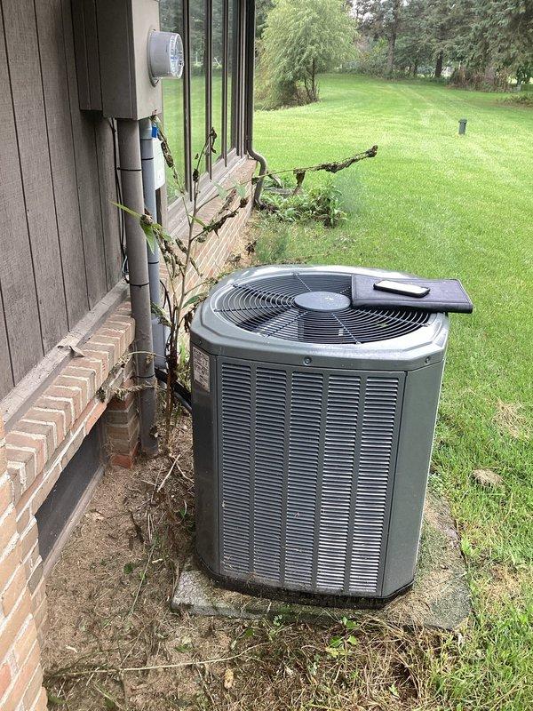 Conducted comprehensive site evaluation of existing outdoor air conditioning system for equipment replacement estimate. Assessed current gray cylindrical condenser unit positioned on concrete pad against residential brick foundation, noting weathered condition with accumulated dirt and grime on surfaces. Documented significant vegetation overgrowth including dead vines and dried plant material encroaching on equipment, which may restrict airflow and reduce system efficiency. Evaluated existing white PVC condensate drainage and refrigerant line covers running vertically along exterior wall. Photographed equipment placement, clearance requirements, and surrounding conditions including concrete pad weathering and neglected maintenance area. Prepared replacement recommendations based on current system configuration, electrical service availability, and proper installation requirements for new equipment.