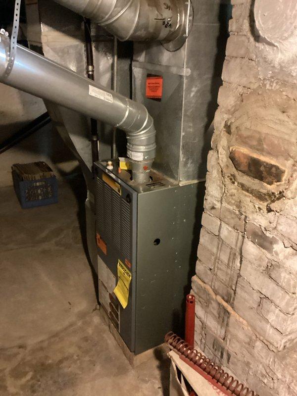 Conducted comprehensive furnace replacement estimate for residential forced-air heating system located in basement mechanical room. Evaluated existing gray cabinet-style furnace unit installed against white brick wall, documenting current equipment specifications, ductwork configuration, and installation conditions. Assessed metallic supply duct connections, ventilation pathways, and clearance requirements for replacement unit sizing and compatibility with existing HVAC infrastructure.