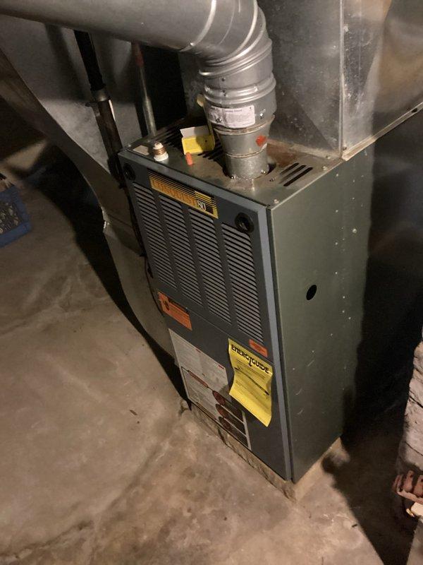 Conducted comprehensive furnace replacement estimate for residential forced-air heating system located in basement mechanical room. Evaluated existing gray cabinet-style furnace unit installed against white brick wall, documenting current equipment specifications, ductwork configuration, and installation conditions. Assessed metallic supply duct connections, ventilation pathways, and clearance requirements for replacement unit sizing and compatibility with existing HVAC infrastructure.
