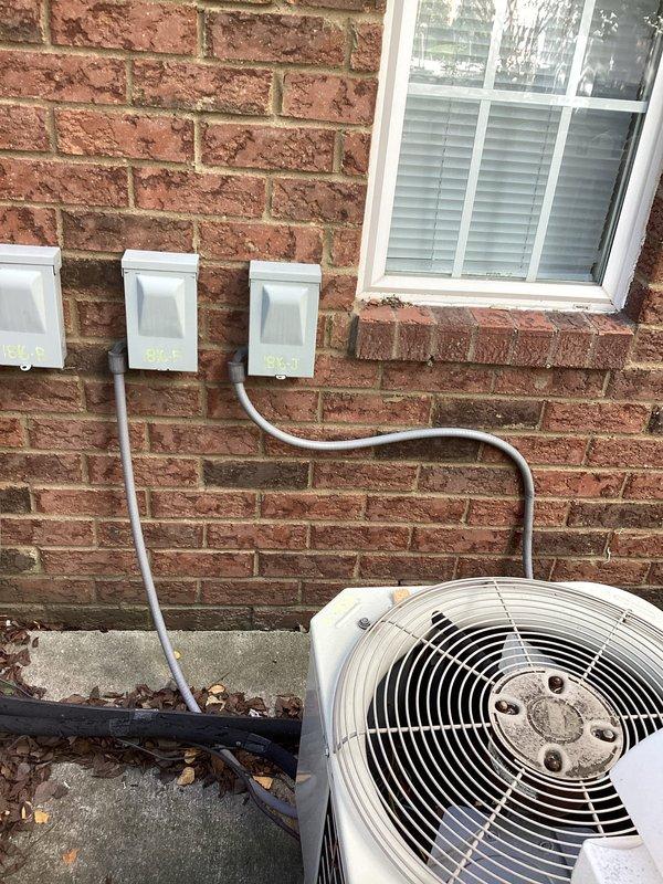 Conducted commercial mini-split system estimate at brick building exterior with existing HVAC equipment. Documented current installation conditions including three wall-mounted electrical junction boxes with conduit connections, existing condenser unit placement on concrete pad, and available service access behind perimeter fencing with mulch landscaping. Assessed wall penetration requirements and evaluated optimal placement for additional mini-split outdoor units adjacent to existing equipment array on concrete mounting surface.