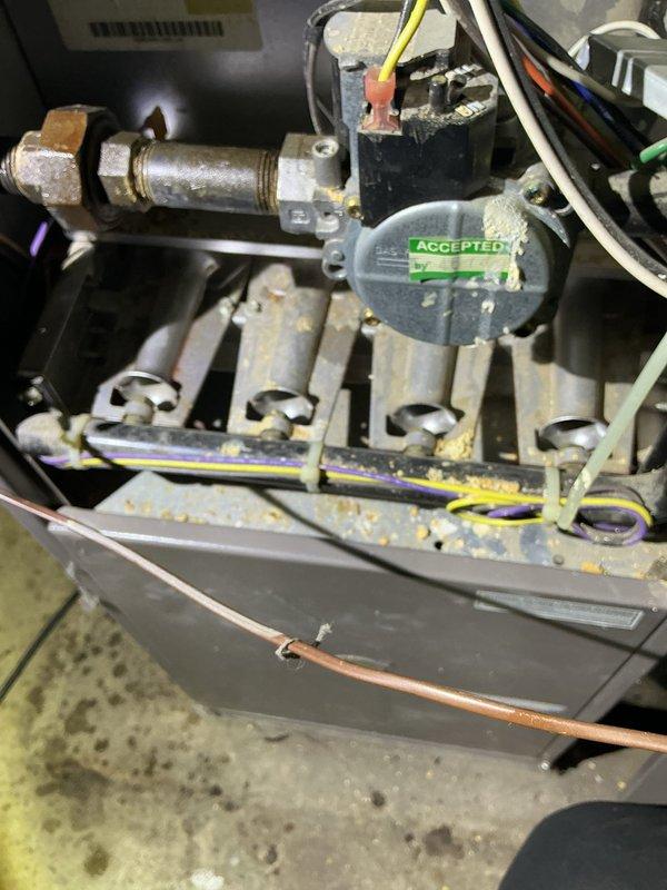 Responded to a gas leak at the furnace and shut off the gas supply to the unit to eliminate the hazard; the gas remains off at this time. Inspection of the gas valve and burner assembly revealed a corroded brass fitting and heavy rust, debris, and residue buildup on the valve, burners, and manifold, so a follow-up visit is required for an HVAC technician to repair or replace the affected components (see attached photos).