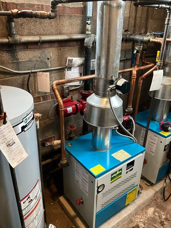 Performed annual boiler tune-up in basement mechanical room, inspecting the blue and white boiler unit with metallic exhaust vent. Checked operation of red circulator pump and verified all copper pipe connections were secure with no leaks present. System functioning properly after service.