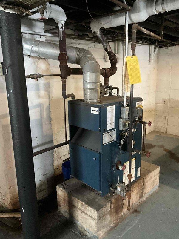 Conducted comprehensive inspection of aging steam heating system, including cast iron radiators showing surface deterioration and rust in residential unit. Examined blue boiler unit mounted on concrete platform with associated piping infrastructure and ventilation components. Documented system condition with photos for assessment and estimate purposes.