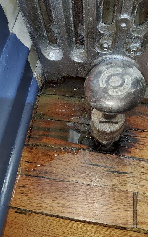 Responded to service call for reported water leakage from radiator system. Upon inspection, identified significant corrosion at pipe connections of cast iron radiator located in dining room. Observed water damage to wooden flooring surrounding radiator pipe penetration and confirmed related ceiling collapse in basement storage area directly below. Documented condition with photographs showing deteriorated fittings and water staining.