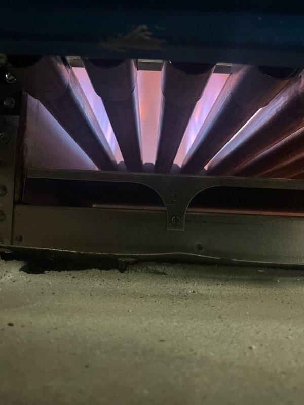 Performed boiler tune-up service, inspecting burner flames which showed proper combustion with characteristic purple-pink coloration. Observed significant oxidation and rust buildup on heat exchanger fins, but system remains operational. Documentation photos taken of burner assembly and heat exchanger condition for service records.