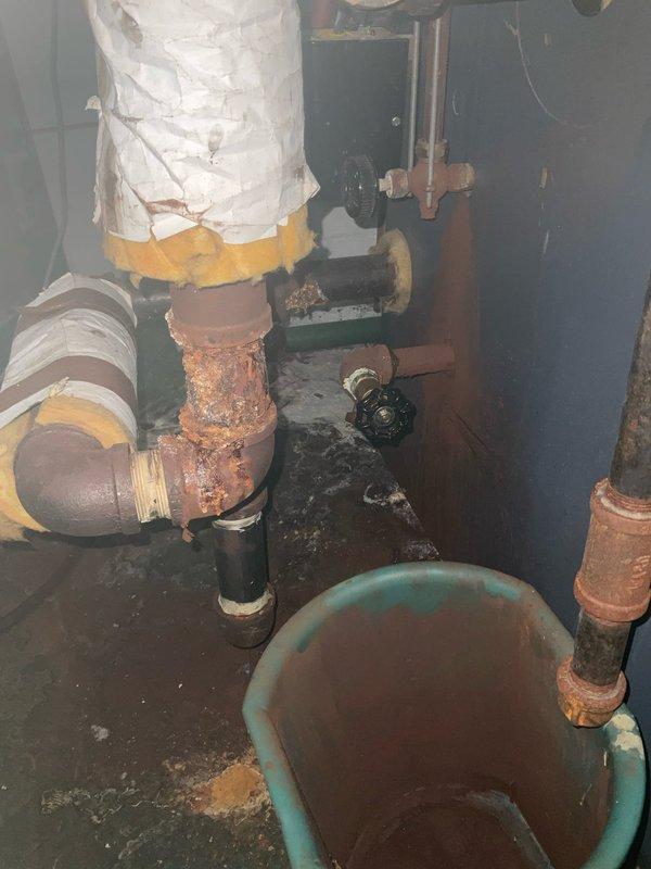 # Service Report: Boiler System Inspection and Diagnosis

Responded to service call for boiler malfunction with customer reporting potential thermostat-related issues. Upon arrival, conducted comprehensive system inspection of aging boiler installation and associated hydronic components.

Visual assessment revealed extensive corrosion throughout the piping system, particularly affecting iron and steel pipe sections. Documented severe oxidation and rust buildup on vertical supply pipes and horizontal return lines, with heavy surface deterioration indicating prolonged exposure to moisture and age-related degradation. Corroded sections exhibited crusty, textured surfaces with significant reddish-brown oxidation that compromised pipe integrity in multiple locations.

Inspected circulator pump assembly and found unit operational but mounted within deteriorated piping infrastructure. Examined electrical connections at pump motor, confirming proper wiring termination at junction box. Evaluate