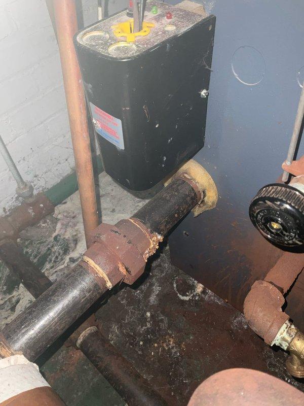 # Service Report: Boiler System Inspection and Diagnosis

Responded to service call for boiler malfunction with customer reporting potential thermostat-related issues. Upon arrival, conducted comprehensive system inspection of aging boiler installation and associated hydronic components.

Visual assessment revealed extensive corrosion throughout the piping system, particularly affecting iron and steel pipe sections. Documented severe oxidation and rust buildup on vertical supply pipes and horizontal return lines, with heavy surface deterioration indicating prolonged exposure to moisture and age-related degradation. Corroded sections exhibited crusty, textured surfaces with significant reddish-brown oxidation that compromised pipe integrity in multiple locations.

Inspected circulator pump assembly and found unit operational but mounted within deteriorated piping infrastructure. Examined electrical connections at pump motor, confirming proper wiring termination at junction box. Evaluate