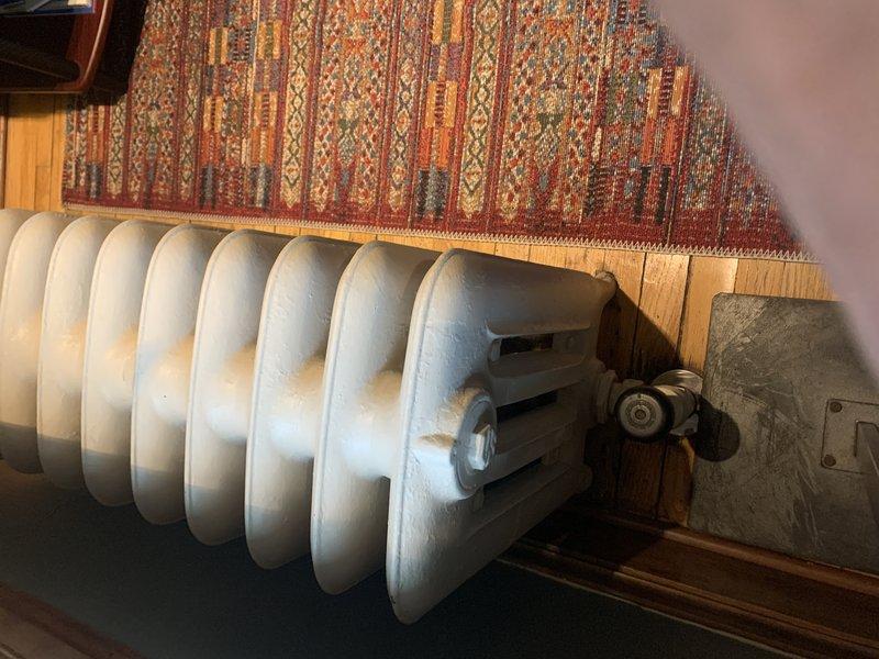 Conducted on-site assessment of residential steam heating system to evaluate cast iron radiator requiring replacement. Inspected vintage multi-column radiator located beneath staircase, documenting existing unit configuration, mounting hardware, and connection points. Evaluated radiator condition noting surface wear and age-related deterioration. Photographed installation area including clearances, piping connections, and surrounding space constraints for accurate replacement specifications. Prepared comprehensive quote for radiator replacement including removal of existing unit and installation of new component compatible with current steam boiler system.