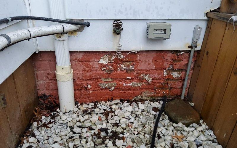 Responded to water leak concern at exterior panels suspected to originate from AC condensate drainage system. Upon inspection, discovered significant water damage and deterioration in basement/crawl space area with compromised wall surfaces, peeling paint, and exposed brickwork. Examined PVC drainage pipes, condensate lines, and associated plumbing infrastructure running along walls and overhead. Documented existing conditions showing white stone drainage bed with debris accumulation, multiple pipe runs including insulated refrigerant lines, copper piping, and flexible conduit installations. Assessed integrity of condensate drain system and mounting hardware. Photographed deteriorated wall conditions, pipe routing, and overhead utility infrastructure for documentation purposes.