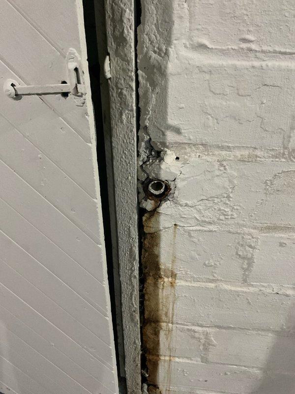 Conducted site evaluation of existing sump pump system in basement utility area to provide estimate for battery backup installation. Assessed current white PVC discharge pipe configuration with vertical run and flexible connector section, noting functional primary pump with black base plate and wall-mounted control components. Identified outdoor spigot requiring permanent capping to prevent water infiltration. Documented significant water damage and staining on interior door and wall surfaces near entry point, indicating historical moisture intrusion issues that necessitate proper exterior penetration sealing when spigot is capped off.