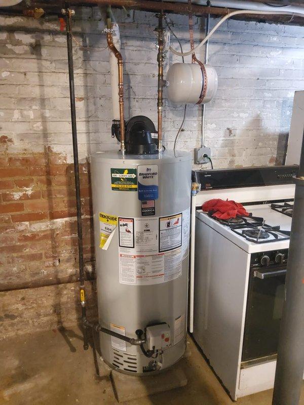 Installed power vent water heater system in commercial building basement utility area. Completed copper piping connections, integrated expansion tank, and routed PVC exhaust vent through exterior wall above brick foundation line with proper elbow configuration for horizontal termination.