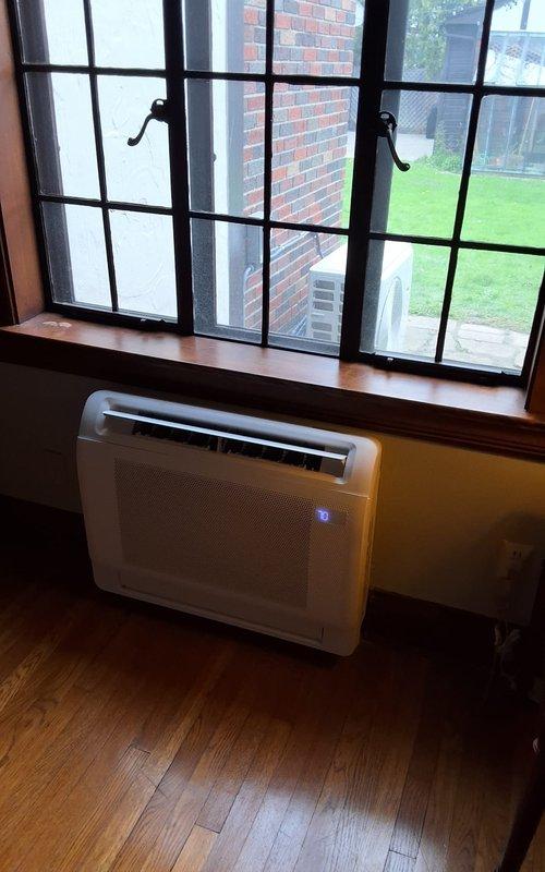 Completed installation of a two-head mini split system with one indoor wall-mounted unit positioned in an interior room beneath a multi-pane window and one outdoor condensing unit mounted on a concrete pad against an exterior brick wall. Refrigerant lines were routed from the condenser through the wall to connect both indoor and outdoor components, electrical disconnect box was installed on the exterior wall for service access, and all connections were tested to ensure proper system operation and climate control functionality.
