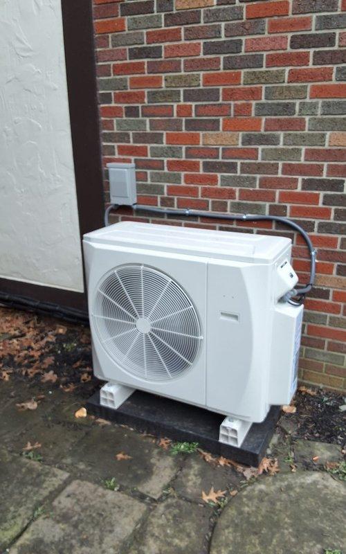 Completed installation of a two-head mini split system with one indoor wall-mounted unit positioned in an interior room beneath a multi-pane window and one outdoor condensing unit mounted on a concrete pad against an exterior brick wall. Refrigerant lines were routed from the condenser through the wall to connect both indoor and outdoor components, electrical disconnect box was installed on the exterior wall for service access, and all connections were tested to ensure proper system operation and climate control functionality.