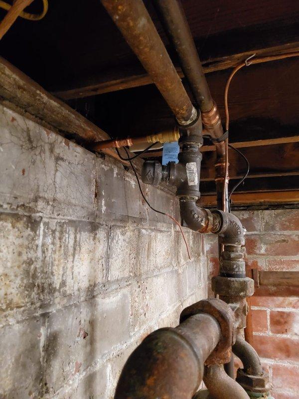 Returned to property for follow-up assessment and quote preparation for boiler piping system repairs and replacement. Evaluated existing aged copper and metal piping infrastructure showing significant oxidation, corrosion, and weathering throughout the assembly. Inspected ghost piping configuration and overall piping system condition in basement area currently undergoing remodeling following recent asbestos abatement. Documented extensive piping network including multiple horizontal and vertical runs, connection joints, valves, and fittings mounted to ceiling joists and wall surfaces. Photographed deteriorated piping conditions, including corroded copper lines with brown and reddish patina, brass fittings, and associated plumbing components. Assessed structural mounting systems and pipe support brackets. Evaluated wall conditions showing moisture damage, efflorescence, and staining patterns that may impact installation planning. Prepared comprehensive visual documentation of existing m