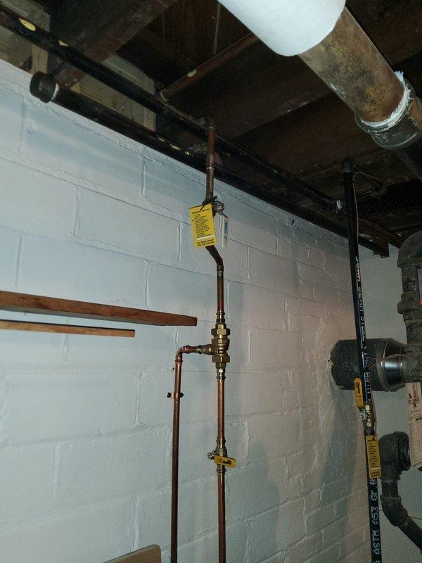 Returned to property for follow-up assessment and quote preparation for boiler piping system repairs and replacement. Evaluated existing aged copper and metal piping infrastructure showing significant oxidation, corrosion, and weathering throughout the assembly. Inspected ghost piping configuration and overall piping system condition in basement area currently undergoing remodeling following recent asbestos abatement. Documented extensive piping network including multiple horizontal and vertical runs, connection joints, valves, and fittings mounted to ceiling joists and wall surfaces. Photographed deteriorated piping conditions, including corroded copper lines with brown and reddish patina, brass fittings, and associated plumbing components. Assessed structural mounting systems and pipe support brackets. Evaluated wall conditions showing moisture damage, efflorescence, and staining patterns that may impact installation planning. Prepared comprehensive visual documentation of existing m