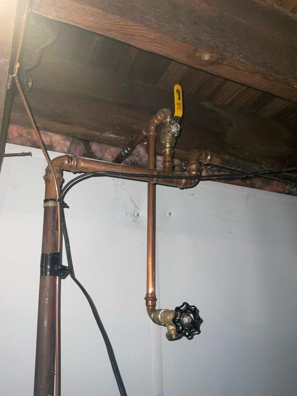 Responded to plumbing service call at century-old residential property for pipe repair assessment. Upon arrival, inspected exposed copper plumbing infrastructure in basement/utility area with accessible ceiling joists and insulation. Evaluated aging copper pipe system showing natural oxidation and patina consistent with older installation. Verified all shut-off valves, including yellow-handled ball valve and black wheel-style valve, were operational and properly functioning. Confirmed water supply remained active with no active leaks or water damage present at time of inspection. Documented existing plumbing configuration including multiple pipe joints, elbows, brass fittings, and connection points throughout the assembly. Assessed overall system integrity and identified areas requiring monitoring due to age-related wear on copper piping.