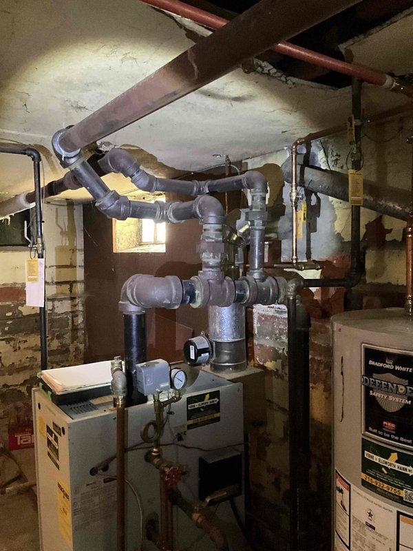 Conducted on-site evaluation of existing steam boiler system in basement mechanical room to provide replacement estimate. Inspected current boiler unit, associated piping configuration including complex PVC and copper distribution lines, and overall HVAC infrastructure. Documented equipment condition, noting age-related wear and system layout. Photographed mechanical room setup, existing boiler assembly, pipe connections, and basement access points to support replacement proposal and equipment sizing requirements.