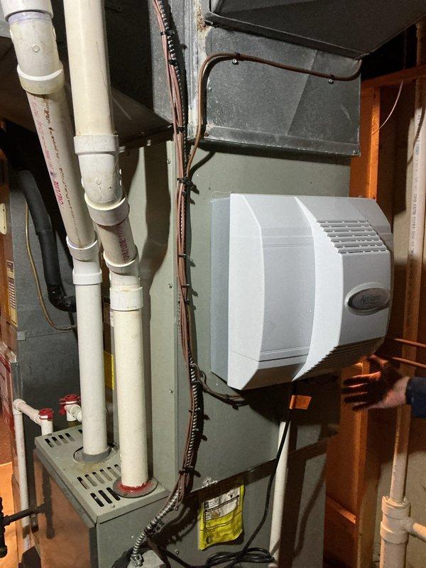 Performed comprehensive evaluation of aging 20+ year old heat pump system during scheduled maintenance appointment. Upon inspection, documented existing HVAC equipment configuration including gray metal furnace/air handler unit with integrated white humidifier assembly, PVC condensate drainage piping, and associated ductwork plenum. System assessment revealed equipment had exceeded its effective service life, prompting recommendation for complete system replacement. Captured detailed photographic documentation of current installation showing furnace unit with manufacturer specification labels, white PVC drain lines with mounting brackets, black electrical conduit, copper refrigerant tubing, and concrete floor installation area. Provided estimate for full HVAC system replacement to address reliability concerns and improve energy efficiency for residential heating and cooling needs.