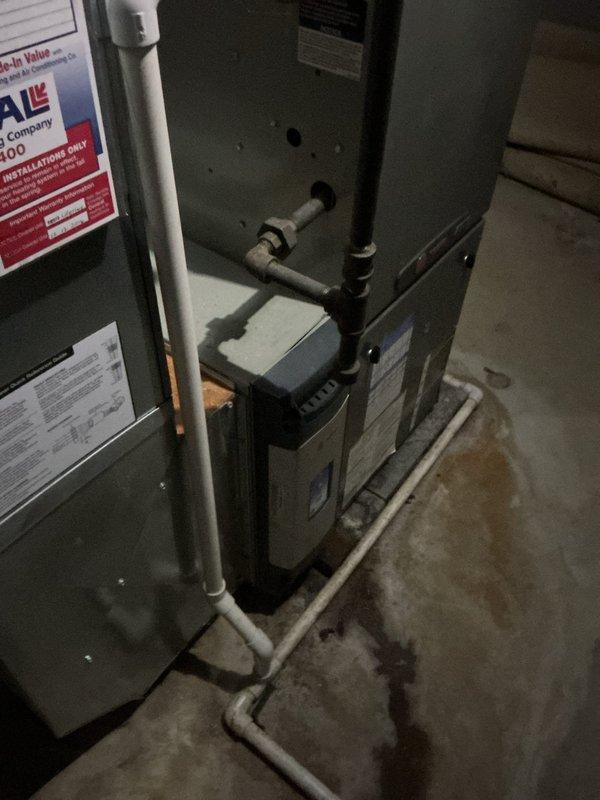 Performed comprehensive evaluation of aging 20+ year old heat pump system during scheduled maintenance appointment. Upon inspection, documented existing HVAC equipment configuration including gray metal furnace/air handler unit with integrated white humidifier assembly, PVC condensate drainage piping, and associated ductwork plenum. System assessment revealed equipment had exceeded its effective service life, prompting recommendation for complete system replacement. Captured detailed photographic documentation of current installation showing furnace unit with manufacturer specification labels, white PVC drain lines with mounting brackets, black electrical conduit, copper refrigerant tubing, and concrete floor installation area. Provided estimate for full HVAC system replacement to address reliability concerns and improve energy efficiency for residential heating and cooling needs.