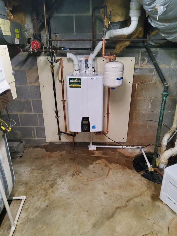 Completed installation of wall-mounted tankless water heater system in basement utility area, including full copper pipe connections, integrated expansion tank, and proper venting with PVC exhaust piping. All plumbing connections were secured to code specifications, unit was mounted on plywood backing board against concrete block wall, and system was tested and confirmed operational.