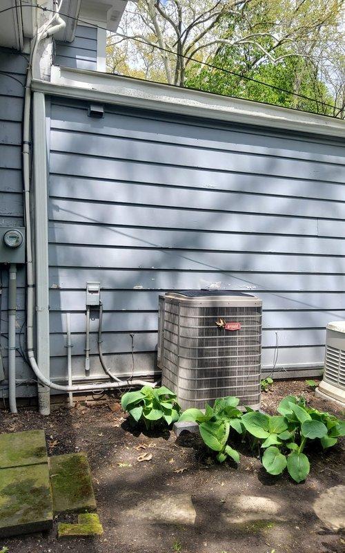Completed preventive maintenance inspection on residential cooling system. Inspected outdoor condenser unit with cylindrical housing, verified proper operation and airflow through mesh grille. Checked electrical disconnect box and associated wiring for secure connections. Examined PVC condensate drain line routing along exterior wall for proper drainage and clearance. Noted concrete pad showing weathering with vegetation growth around perimeter; unit remains level and stable on pad.

Proceeded to basement mechanical room to inspect indoor air handling components and associated plumbing. Verified operation of shutoff valves including red gate valve on water supply lines. Inspected copper and steel piping for leaks, corrosion, and proper support. Noted age-related patina on copper lines but found no active leaks or structural concerns. System operating within normal parameters. All safety controls tested and functioning properly.