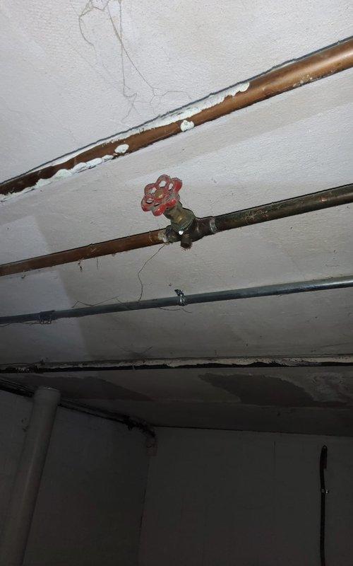 Completed preventive maintenance inspection on residential cooling system. Inspected outdoor condenser unit with cylindrical housing, verified proper operation and airflow through mesh grille. Checked electrical disconnect box and associated wiring for secure connections. Examined PVC condensate drain line routing along exterior wall for proper drainage and clearance. Noted concrete pad showing weathering with vegetation growth around perimeter; unit remains level and stable on pad.

Proceeded to basement mechanical room to inspect indoor air handling components and associated plumbing. Verified operation of shutoff valves including red gate valve on water supply lines. Inspected copper and steel piping for leaks, corrosion, and proper support. Noted age-related patina on copper lines but found no active leaks or structural concerns. System operating within normal parameters. All safety controls tested and functioning properly.