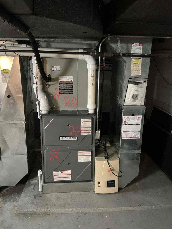 Conducted on-site evaluation of 13-year-old residential HVAC system for replacement estimate. Assessed outdoor condensing unit showing significant age-related deterioration, including rust streaking, corrosion around mounting hardware, and weathered housing. Noted unit installed on ground with leaf debris and vegetation growth rather than proper concrete pad. Inspected indoor equipment including furnace and air handler in basement location. Documented existing system configuration with connected ductwork and control components. Photographed equipment condition and installation details to support replacement recommendation.