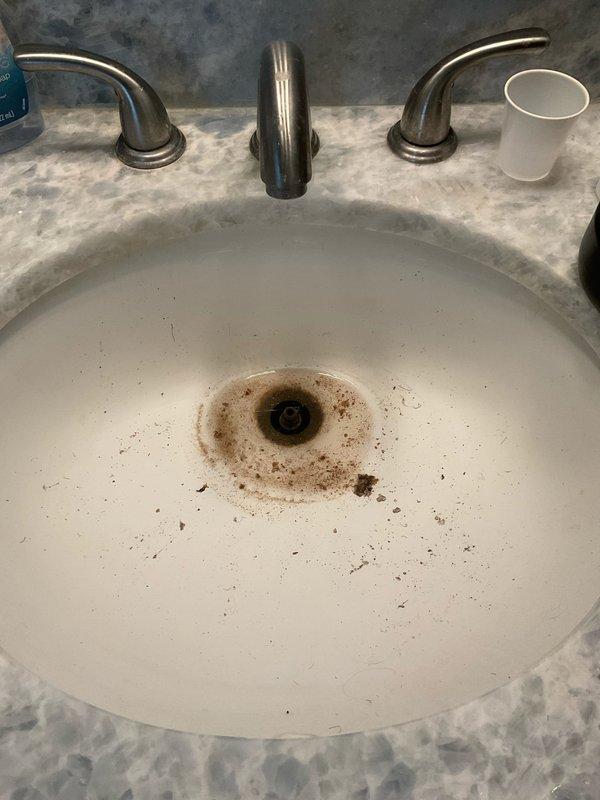 Responded to clogged drain issue in bathroom sink of century-old home with recent remodel. Upon inspection, identified blockage in drain line and observed rust-colored staining around drain opening indicating possible mineral buildup or deteriorating components. Accessed P-trap assembly beneath sink through vanity cabinet, which featured white PVC slip-joint configuration connected to vertical drain pipe. Disassembled P-trap sections to clear obstruction and remove accumulated debris. Inspected all drain components for proper alignment and secure connections. Cleaned trap sections and reassembled plumbing system, ensuring all slip-joint fittings were properly tightened to prevent future leaks. Tested drain flow with running water to verify complete clearance and proper drainage. System restored to full functionality with no leaks detected at connections.