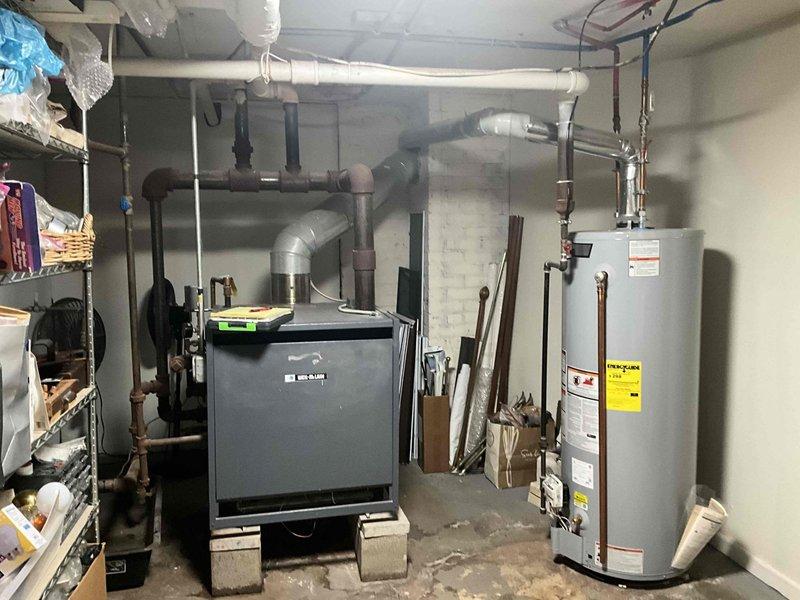 Responded to service call for boiler leaking hot water from bottom, upper left-hand corner. Upon arrival, parked in back of driveway per customer request and proceeded to basement mechanical room to inspect the heating system. Located gray metal boiler unit elevated on concrete blocks with dual exhaust pipes and associated plumbing connections. Conducted thorough visual inspection of the boiler assembly, examining all pipe connections, valves, and fittings in the affected area. Documented equipment condition through photographs showing the boiler installation, surrounding piping infrastructure including copper and PVC lines, and overall mechanical room layout. Assessed leak source and severity to determine appropriate repair recommendations and provided estimate for necessary corrective action to resolve the hot water leak issue.