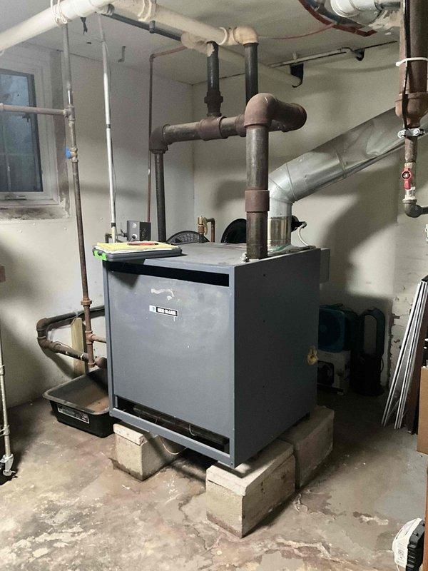 Responded to service call for boiler leaking hot water from bottom, upper left-hand corner. Upon arrival, parked in back of driveway per customer request and proceeded to basement mechanical room to inspect the heating system. Located gray metal boiler unit elevated on concrete blocks with dual exhaust pipes and associated plumbing connections. Conducted thorough visual inspection of the boiler assembly, examining all pipe connections, valves, and fittings in the affected area. Documented equipment condition through photographs showing the boiler installation, surrounding piping infrastructure including copper and PVC lines, and overall mechanical room layout. Assessed leak source and severity to determine appropriate repair recommendations and provided estimate for necessary corrective action to resolve the hot water leak issue.