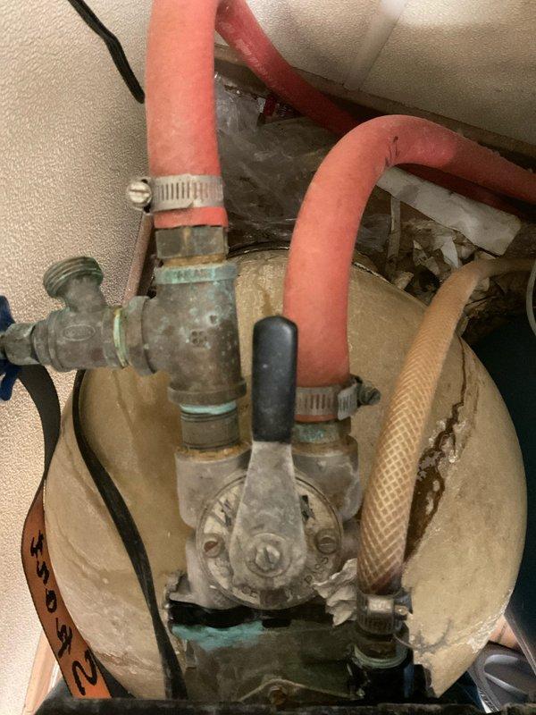 Responded to service call for leaking water softener unit located in meat room. Upon arrival, inspected the system and identified active leak at valve assembly connections. Documented equipment condition showing visible corrosion, mineral deposits, and aging components on brass fittings. Assessed flexible hose connections and clamps for integrity. Photographed rental unit tag information and internal plumbing configuration including corroded valve mechanism, flexible supply lines, and corrugated hoses for service records and potential replacement recommendations.